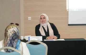 First image shows a woman in a light-colored headscarf and dark clothing seated at a table with papers and a name tag reading something like a participants name in a conference room with wooden paneling a projector screen and chairs. Second image depicts a diverse group of attendees including men and women some wearing headscarves in a warmly lit room with wooden walls seated on chairs facing forward during a session. Third image captures a conference setup with a woman in a headscarf standing at a podium next to a laptop and projector screen displaying slides tables with documents and audience members in chairs. Fourth image features a panel discussion area with a man at a desk in front of a screen showing an image a Turkish flag on the wall banners and attendees seated in chairs in a room with wooden walls and lighting.