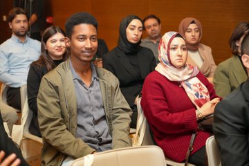 First image shows a woman in a light-colored headscarf and dark clothing seated at a table with papers and a name tag reading something like a participants name in a conference room with wooden paneling a projector screen and chairs. Second image depicts a diverse group of attendees including men and women some wearing headscarves in a warmly lit room with wooden walls seated on chairs facing forward during a session. Third image captures a conference setup with a woman in a headscarf standing at a podium next to a laptop and projector screen displaying slides tables with documents and audience members in chairs. Fourth image features a panel discussion area with a man at a desk in front of a screen showing an image a Turkish flag on the wall banners and attendees seated in chairs in a room with wooden walls and lighting.