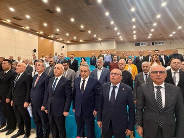 First image shows a man in a dark suit and tie standing at a wooden podium with microphones, gesturing with both hands outstretched, in front of a light wooden wall and two Turkish flags on poles. Second image depicts a conference room with a projection screen displaying a title FARKLILIK CUNKU and images of documents and a logo, three men in suits seated at a table with microphones, Turkish flags and banners for Turk Buro-Sen and Turkiye Kamu-Sen on the table and walls, a large portrait of Mustafa Kemal Ataturk. Third image features a modern building projected on a screen behind a podium, several men in suits seated and one speaking at the podium with a microphone, banners with eye symbols and Turkish flags on wooden tables. Fourth image shows a large group of about 30 people mostly men in suits standing in rows on a stage with blue backdrop, some holding documents, in a formal assembly setting.