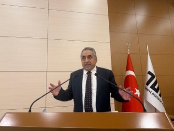 First image shows a man in a dark suit and tie standing at a wooden podium with microphones, gesturing with both hands outstretched, in front of a light wooden wall and two Turkish flags on poles. Second image depicts a conference room with a projection screen displaying a title FARKLILIK CUNKU and images of documents and a logo, three men in suits seated at a table with microphones, Turkish flags and banners for Turk Buro-Sen and Turkiye Kamu-Sen on the table and walls, a large portrait of Mustafa Kemal Ataturk. Third image features a modern building projected on a screen behind a podium, several men in suits seated and one speaking at the podium with a microphone, banners with eye symbols and Turkish flags on wooden tables. Fourth image shows a large group of about 30 people mostly men in suits standing in rows on a stage with blue backdrop, some holding documents, in a formal assembly setting.