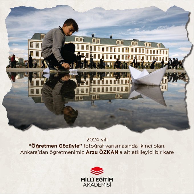 A young boy with short dark hair wearing a gray hooded sweatshirt and dark pants kneels on a reflective wet pavement surface outdoors. He holds and folds a white paper boat in his hands. In the background a large pale yellow multi-story historic building with many rectangular windows and arched doorways stands under a sky with scattered clouds. Several people are visible in the distance near the building. The wet ground mirrors the building boy and people creating a symmetrical reflection effect. The scene appears calm and educational focused on the boys activity.