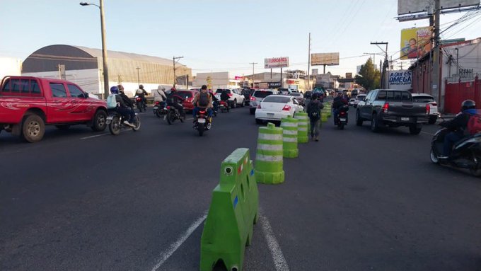 Urban street scene during daylight with multiple vehicles including red pickup trucks, white sedans, and motorcycles navigating around green and white striped traffic cones on a divided road. Billboards and a large warehouse structure appear in the background under a clear sky. Motorcyclists in helmets are visible among the cars in the congested traffic flow.