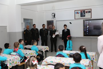 First image shows group of military officers in uniforms standing with many young students wearing blue tracksuits and holding Turkish flags in front of school building with Ayvalıdere Primary School sign and Turkish flag. Second image depicts officers presenting flowers to students at desks in classroom with smartboard and portraits on walls. Third image captures children in blue outfits engaged in craft activity around tables with red white decorations and Turkish flag banner in classroom. Fourth image features officers distributing gifts to smiling children at tables with red white balloon decorations and school banner.