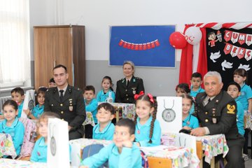 First image shows group of military officers in uniforms standing with many young students wearing blue tracksuits and holding Turkish flags in front of school building with Ayvalıdere Primary School sign and Turkish flag. Second image depicts officers presenting flowers to students at desks in classroom with smartboard and portraits on walls. Third image captures children in blue outfits engaged in craft activity around tables with red white decorations and Turkish flag banner in classroom. Fourth image features officers distributing gifts to smiling children at tables with red white balloon decorations and school banner.
