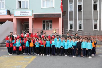 First image shows group of military officers in uniforms standing with many young students wearing blue tracksuits and holding Turkish flags in front of school building with Ayvalıdere Primary School sign and Turkish flag. Second image depicts officers presenting flowers to students at desks in classroom with smartboard and portraits on walls. Third image captures children in blue outfits engaged in craft activity around tables with red white decorations and Turkish flag banner in classroom. Fourth image features officers distributing gifts to smiling children at tables with red white balloon decorations and school banner.