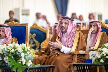 Multiple images depict Saudi men in traditional white thobes and red checkered headscarves seated in ornate blue and gold chairs during a formal event. One image shows a man in brown robe gesturing with hands clasped near flower arrangements on wooden tables. Another focuses on a smiling man in red and white headscarf leaning forward in discussion. A group photo captures attendees clapping in a decorated hall with blue drapes and a speaker setup. The final image features a man in brown robe standing beside a white plaque with Arabic text and a camel emblem on a pedestal against a blue backdrop.