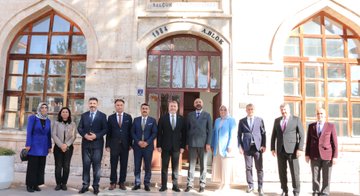 First image shows a group of adults seated in a modern room with white walls, plants, and educational posters, some wearing suits and headscarves, engaged in discussion around low tables with notebooks and bags. Second image depicts men in formal suits and jackets sitting around an oval wooden table with documents and a phone, one man gesturing while speaking. Third image captures a seminar in a wood-paneled auditorium with red seats, a screen displaying Turkish flag and text, speakers at a podium, and audience members including women in headscarves listening attentively. Fourth image features a group of about ten adults, men in suits and women in headscarves and coats, standing in a line in front of a stone building with arched windows and a sign reading 1864 above the entrance.
