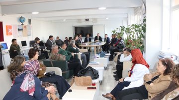 First image shows a group of adults seated in a modern room with white walls, plants, and educational posters, some wearing suits and headscarves, engaged in discussion around low tables with notebooks and bags. Second image depicts men in formal suits and jackets sitting around an oval wooden table with documents and a phone, one man gesturing while speaking. Third image captures a seminar in a wood-paneled auditorium with red seats, a screen displaying Turkish flag and text, speakers at a podium, and audience members including women in headscarves listening attentively. Fourth image features a group of about ten adults, men in suits and women in headscarves and coats, standing in a line in front of a stone building with arched windows and a sign reading 1864 above the entrance.