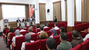 First image shows a group of adults seated in a modern room with white walls, plants, and educational posters, some wearing suits and headscarves, engaged in discussion around low tables with notebooks and bags. Second image depicts men in formal suits and jackets sitting around an oval wooden table with documents and a phone, one man gesturing while speaking. Third image captures a seminar in a wood-paneled auditorium with red seats, a screen displaying Turkish flag and text, speakers at a podium, and audience members including women in headscarves listening attentively. Fourth image features a group of about ten adults, men in suits and women in headscarves and coats, standing in a line in front of a stone building with arched windows and a sign reading 1864 above the entrance.