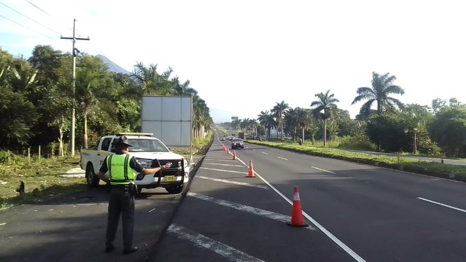 First image shows a white Toyota Hilux pickup truck marked with PROVIAL and PROTEGER Y SERVIR logos parked on a multi-lane highway at km 56 with orange traffic cones placed around it palm trees lining the roadside a green sign indicating Escuintla and approaching red vehicles under a partly cloudy sky. Second image depicts a uniformed officer in a high-visibility yellow vest standing beside the same white PROVIAL-marked truck on a straight highway with additional orange cones setting up a speed reduction point surrounded by lush green vegetation a distant mountain and utility poles with wires.