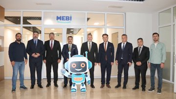 First image shows eight men in suits and casual attire standing in a modern office entrance with MEBİ logo on the wall and a large blue robot mascot figure waving nearby. Second image depicts over ten men in formal suits gathered around a desk and whiteboard in a classroom-like room with wooden cabinets and a projector screen. Third image features five men, one gesturing while standing near desks with laptops, monitors, and office chairs in a workspace. Fourth image displays nine men in mixed attire including suits and sweaters positioned around desks in an open office area with electrical outlets visible.