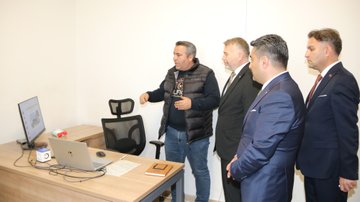 First image shows eight men in suits and casual attire standing in a modern office entrance with MEBİ logo on the wall and a large blue robot mascot figure waving nearby. Second image depicts over ten men in formal suits gathered around a desk and whiteboard in a classroom-like room with wooden cabinets and a projector screen. Third image features five men, one gesturing while standing near desks with laptops, monitors, and office chairs in a workspace. Fourth image displays nine men in mixed attire including suits and sweaters positioned around desks in an open office area with electrical outlets visible.