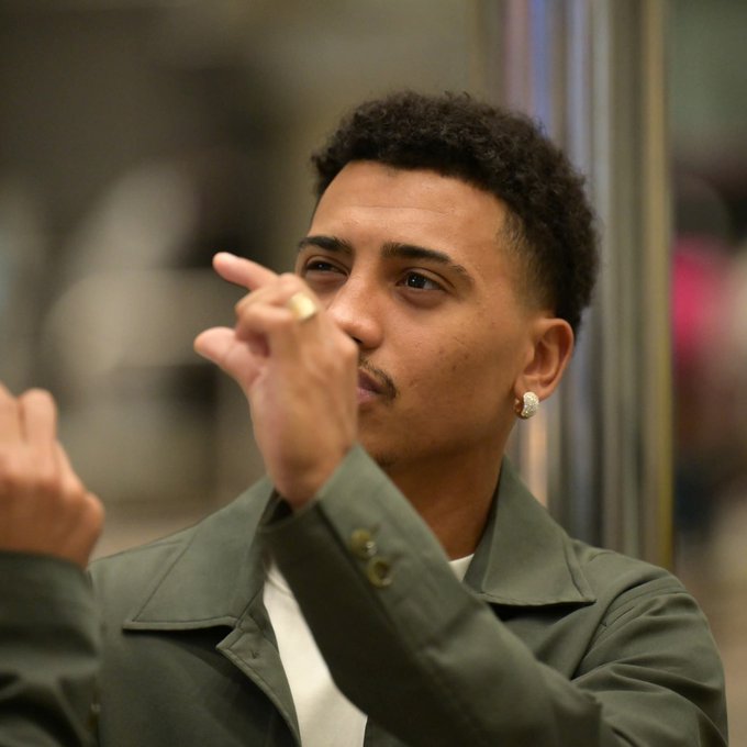 A young man with short curly black hair, wearing a green jacket over a white shirt, points with his index finger while holding a lit cigarette near his mouth, gold ring on finger, gold hoop earring in one ear, standing in front of a reflective surface with blurred pink and neutral background elements.