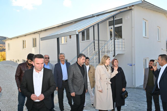 First image shows a group of eight formally dressed men and women standing on steps in front of a white arched building with blue Islamic decorative tiles, lanterns, and Arabic script plaques, featuring wooden double doors and columns, set against a mountainous landscape. Second image depicts a larger group of about fifteen people in suits, coats, and traditional attire gathered on gravel near a modern white building with a sloped roof and metal stairs, some holding bags, with construction materials like gravel piles visible nearby and hills in the background.