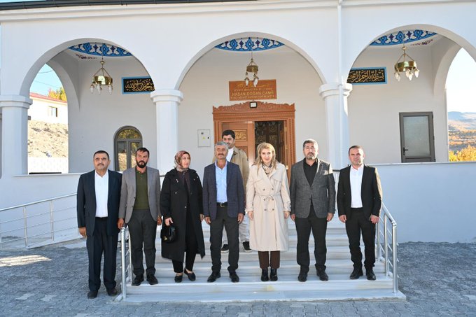 First image shows a group of eight formally dressed men and women standing on steps in front of a white arched building with blue Islamic decorative tiles, lanterns, and Arabic script plaques, featuring wooden double doors and columns, set against a mountainous landscape. Second image depicts a larger group of about fifteen people in suits, coats, and traditional attire gathered on gravel near a modern white building with a sloped roof and metal stairs, some holding bags, with construction materials like gravel piles visible nearby and hills in the background.