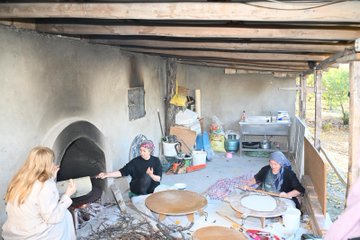 First image shows two women one in light coat and brown pants holding a tool with flatbread over a curved stone oven with fire and wood below seated on pink cushion with black stool nearby. Second image depicts a blonde woman in beige coat and brown pants holding large flatbread near red plastic chair and blue hat with scattered wood and ash on floor. Third image captures three women in a mud-walled room with oven one blonde woman extending tool to oven two others in headscarves preparing dough on round wooden tables with buckets and fabrics around. Fourth image features two women one in coat gesturing near oven with flatbread being handled by woman in black shirt headscarf on pink stool with wood piles and round dough tray.