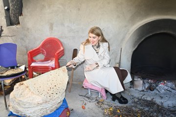First image shows two women one in light coat and brown pants holding a tool with flatbread over a curved stone oven with fire and wood below seated on pink cushion with black stool nearby. Second image depicts a blonde woman in beige coat and brown pants holding large flatbread near red plastic chair and blue hat with scattered wood and ash on floor. Third image captures three women in a mud-walled room with oven one blonde woman extending tool to oven two others in headscarves preparing dough on round wooden tables with buckets and fabrics around. Fourth image features two women one in coat gesturing near oven with flatbread being handled by woman in black shirt headscarf on pink stool with wood piles and round dough tray.