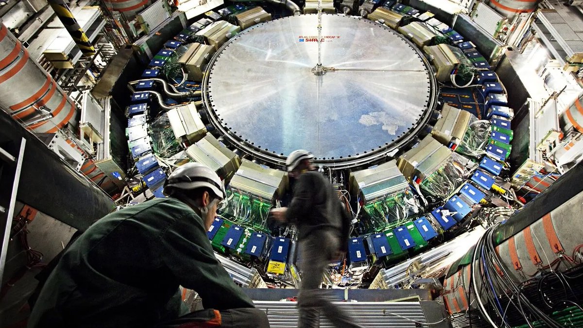 Two workers wearing green overalls and blue hard hats stand and move around the base of a large circular metallic structure with blue and white panels, numerous cables, and piping extending from it. The structure appears to be the Large Hadron Collider particle accelerator in a vast underground facility. Surrounding elements include railings, control panels, and industrial equipment visible in the background.
