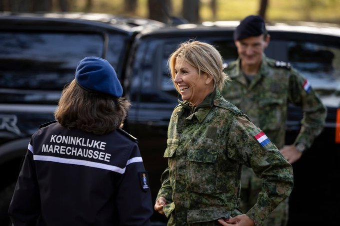 Queen Maxima wearing a blue beret and dark uniform with Dutch insignia shakes hands with a soldier in green camouflage helmet and vest in a forested setting surrounded by multiple troops in similar attire some with rifles and Dutch flags on shoulders. Another image shows her from behind conversing with a smiling female soldier in camouflage next to black vehicles labeled Koninklijke Marechaussee with a male officer nearby. A third image captures soldiers in prone positions with rifles aimed forward in woodland terrain under tree cover. The fourth image features two soldiers in camouflage helmets and vests standing amid trees with gear and rifles in a wooded exercise area.