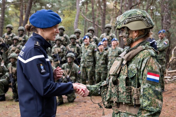 Queen Maxima wearing a blue beret and dark uniform with Dutch insignia shakes hands with a soldier in green camouflage helmet and vest in a forested setting surrounded by multiple troops in similar attire some with rifles and Dutch flags on shoulders. Another image shows her from behind conversing with a smiling female soldier in camouflage next to black vehicles labeled Koninklijke Marechaussee with a male officer nearby. A third image captures soldiers in prone positions with rifles aimed forward in woodland terrain under tree cover. The fourth image features two soldiers in camouflage helmets and vests standing amid trees with gear and rifles in a wooded exercise area.