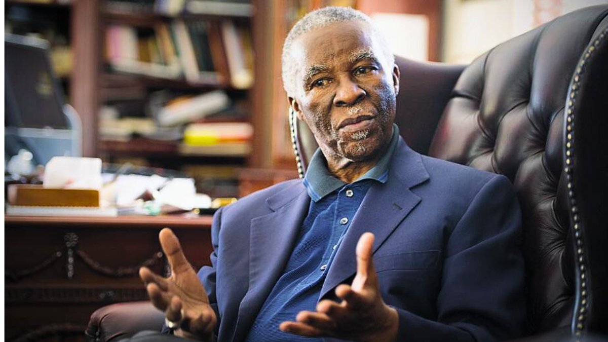 A portrait of an elderly Black man with gray hair and a beard, likely Thabo Mbeki, seated in a dark leather armchair in a wood-paneled study filled with bookshelves and scattered papers; he is wearing a light blue collared shirt under a dark blue suit jacket, gesturing openly with both hands as if speaking emphatically, with a thoughtful expression.