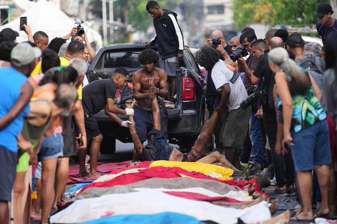 Crowd of police officers in uniforms and journalists with cameras gather around several bodies covered in red yellow and white fabrics and plastic sheets laid out on the asphalt near a black SUV vehicle. People including men in casual clothing and women in shorts assist in lifting and loading the covered bodies into the vehicle. The scene occurs on a street with green trees and urban buildings in the background under daylight.