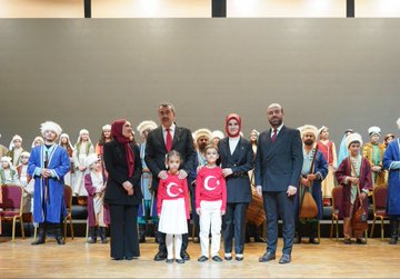 First image shows group of people including men in suits and traditional costumes women in headscarves and dresses children in red shirts with Turkish flag emblem standing on wooden stage with chairs and instruments like drums in background gray walls. Second image depicts officials in formal attire including man in black suit with red tie woman in white top and black skirt holding Turkish flags audience seated waving flags in hall with blue seats and wall lights. Third image captures seated audience and officials in suits some holding Turkish flags in auditorium with blue seats and stage table with glasses and flowers. Fourth image displays large group in colorful traditional outfits like robes and headpieces sitting on stage with large screen showing tree and rider silhouette in sunset hues.