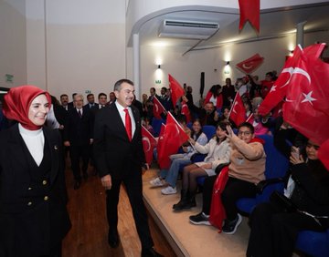 First image shows group of people including men in suits and traditional costumes women in headscarves and dresses children in red shirts with Turkish flag emblem standing on wooden stage with chairs and instruments like drums in background gray walls. Second image depicts officials in formal attire including man in black suit with red tie woman in white top and black skirt holding Turkish flags audience seated waving flags in hall with blue seats and wall lights. Third image captures seated audience and officials in suits some holding Turkish flags in auditorium with blue seats and stage table with glasses and flowers. Fourth image displays large group in colorful traditional outfits like robes and headpieces sitting on stage with large screen showing tree and rider silhouette in sunset hues.