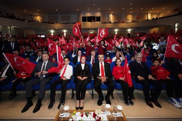 First image shows group of people including men in suits and traditional costumes women in headscarves and dresses children in red shirts with Turkish flag emblem standing on wooden stage with chairs and instruments like drums in background gray walls. Second image depicts officials in formal attire including man in black suit with red tie woman in white top and black skirt holding Turkish flags audience seated waving flags in hall with blue seats and wall lights. Third image captures seated audience and officials in suits some holding Turkish flags in auditorium with blue seats and stage table with glasses and flowers. Fourth image displays large group in colorful traditional outfits like robes and headpieces sitting on stage with large screen showing tree and rider silhouette in sunset hues.