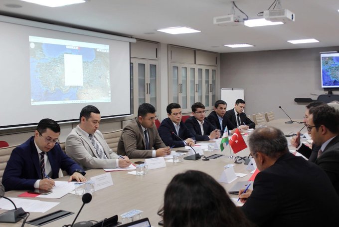 First image shows a group of men in suits seated around a long conference table in a modern room with gray walls and a large screen displaying a map in the background. Turkish and Uzbek flags are placed on the table along with documents laptops water glasses and microphones. Participants appear engaged in discussion some leaning forward with papers. Second image depicts another meeting scene with men and one woman in professional attire at a wooden table under bright lighting with a projector screen showing maps. Greek flag Turkish flag and another flag are visible on the table amid documents glasses and a laptop. Attendees are focused on conversation with some writing notes.