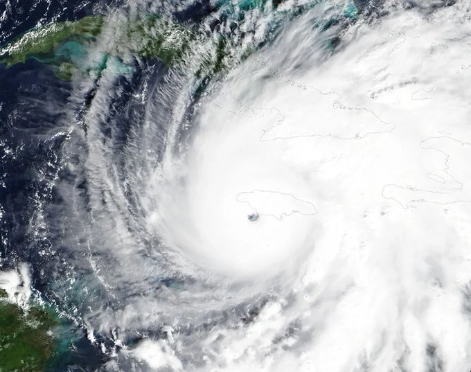 Satellite image depicts a large swirling hurricane cloud formation centered over the island of Cuba with the eye directly above the landmass surrounded by turbulent white clouds and dark ocean waters to the north and east lighter green and blue hues indicating coastal areas and the Caribbean Sea extending southward