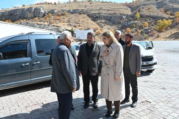 First image shows a group of people, including women in headscarves and men in suits, seated around a long table with red edges in a room with white curtains and wooden cabinets, clapping hands together. Bowls and a teapot are on the table, and some hold prayer beads. Second image depicts a similar group seated around the same table, with individuals in formal attire engaged in conversation or gestures, surrounded by household items like bottles on shelves. Third image features several people, including elderly men in suits and a blonde woman in a beige coat, standing and talking outdoors near gray vans on a paved area with rocky hills and autumn trees in the background.