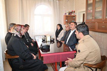 First image shows a group of people, including women in headscarves and men in suits, seated around a long table with red edges in a room with white curtains and wooden cabinets, clapping hands together. Bowls and a teapot are on the table, and some hold prayer beads. Second image depicts a similar group seated around the same table, with individuals in formal attire engaged in conversation or gestures, surrounded by household items like bottles on shelves. Third image features several people, including elderly men in suits and a blonde woman in a beige coat, standing and talking outdoors near gray vans on a paved area with rocky hills and autumn trees in the background.