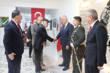 Multiple images depict formal handshakes during a ceremony in a room with Turkish flags and a wall featuring Turkish commemorative text and silhouette. Dignitaries including men in suits and military uniforms with medals and swords exchange greetings. A woman in red attire participates in one handshake. The setting includes decorative lighting and official plaques.