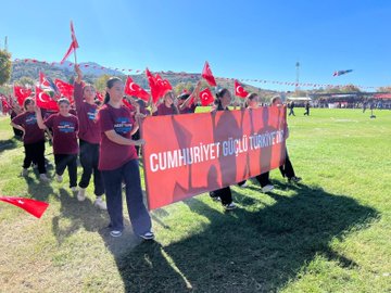 First image shows a line of people mostly in black clothing holding red Turkish flags with white crescents and stars on a grassy field under blue sky with a tall structure in background and Turkish flag garlands. Second image depicts a group of people in maroon shirts carrying a large red banner reading Cumhuriyeti Türkiye with Turkish flags around on grass under sunny sky with mountains. Third image from above shows people arranged in formation creating a large white crescent and star on green grass with a portrait of a man in suit and Turkish flag nearby. Fourth image features young children in black shirts holding small Turkish flags walking on grass with more flags and people in background.