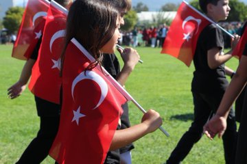 First image shows a line of people mostly in black clothing holding red Turkish flags with white crescents and stars on a grassy field under blue sky with a tall structure in background and Turkish flag garlands. Second image depicts a group of people in maroon shirts carrying a large red banner reading Cumhuriyeti Türkiye with Turkish flags around on grass under sunny sky with mountains. Third image from above shows people arranged in formation creating a large white crescent and star on green grass with a portrait of a man in suit and Turkish flag nearby. Fourth image features young children in black shirts holding small Turkish flags walking on grass with more flags and people in background.