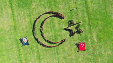 First image shows a line of people mostly in black clothing holding red Turkish flags with white crescents and stars on a grassy field under blue sky with a tall structure in background and Turkish flag garlands. Second image depicts a group of people in maroon shirts carrying a large red banner reading Cumhuriyeti Türkiye with Turkish flags around on grass under sunny sky with mountains. Third image from above shows people arranged in formation creating a large white crescent and star on green grass with a portrait of a man in suit and Turkish flag nearby. Fourth image features young children in black shirts holding small Turkish flags walking on grass with more flags and people in background.