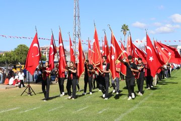 First image shows a line of people mostly in black clothing holding red Turkish flags with white crescents and stars on a grassy field under blue sky with a tall structure in background and Turkish flag garlands. Second image depicts a group of people in maroon shirts carrying a large red banner reading Cumhuriyeti Türkiye with Turkish flags around on grass under sunny sky with mountains. Third image from above shows people arranged in formation creating a large white crescent and star on green grass with a portrait of a man in suit and Turkish flag nearby. Fourth image features young children in black shirts holding small Turkish flags walking on grass with more flags and people in background.