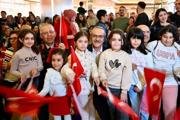 First image shows a diverse group of children and adults standing together in a modern hall many holding red Turkish flags with white crescents and stars some children wearing casual sweaters with English text like CHIC and USA others in traditional or formal attire background includes blurred crowd and architectural elements like large windows and pillars. Second image captures a close-up of an elderly man in glasses and suit jacket embracing a young girl in white dress and red skirt both smiling while holding a Turkish flag nearby people in military uniform and civilian clothes also wave flags seated arrangement suggests an official gathering. Third image features several young girls in white dresses and red skirts standing around middle-aged men in suits one man with glasses holds a girl others wave Turkish flags setting is a curved indoor space with hanging decorations and audience in background. Fourth image displays a seated audience including men in suits and military uniforms children on laps holding flags women in headscarves and casual outfits all waving Turkish flags in a large auditorium-like venue with overhead lighting and banners.