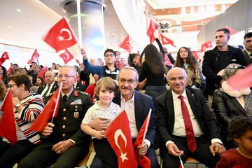 First image shows a diverse group of children and adults standing together in a modern hall many holding red Turkish flags with white crescents and stars some children wearing casual sweaters with English text like CHIC and USA others in traditional or formal attire background includes blurred crowd and architectural elements like large windows and pillars. Second image captures a close-up of an elderly man in glasses and suit jacket embracing a young girl in white dress and red skirt both smiling while holding a Turkish flag nearby people in military uniform and civilian clothes also wave flags seated arrangement suggests an official gathering. Third image features several young girls in white dresses and red skirts standing around middle-aged men in suits one man with glasses holds a girl others wave Turkish flags setting is a curved indoor space with hanging decorations and audience in background. Fourth image displays a seated audience including men in suits and military uniforms children on laps holding flags women in headscarves and casual outfits all waving Turkish flags in a large auditorium-like venue with overhead lighting and banners.