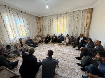 First image shows a group of about 15-20 people mostly men in suits and traditional attire with some women seated on floor cushions around a patterned rug in a room with beige curtains and wooden furniture one man in blue shirt gestures with hand raised during discussion. Second image depicts similar group of men and women in formal clothing sitting on cushions on a rug in a light-filled room with white curtains and a lamp some hold notebooks one man in gray suit points forward. Third image features a group of around 10 people including men in suits and a blonde woman in beige trench coat standing outdoors near white vehicles and tents on gravel under cloudy sky with autumn trees some smile and gesture. Fourth image shows similar outdoor group with men in various coats and the blonde woman in trench coat standing together near cars and tents on dirt ground engaging in conversation.