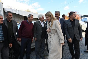 First image shows a group of about 15-20 people mostly men in suits and traditional attire with some women seated on floor cushions around a patterned rug in a room with beige curtains and wooden furniture one man in blue shirt gestures with hand raised during discussion. Second image depicts similar group of men and women in formal clothing sitting on cushions on a rug in a light-filled room with white curtains and a lamp some hold notebooks one man in gray suit points forward. Third image features a group of around 10 people including men in suits and a blonde woman in beige trench coat standing outdoors near white vehicles and tents on gravel under cloudy sky with autumn trees some smile and gesture. Fourth image shows similar outdoor group with men in various coats and the blonde woman in trench coat standing together near cars and tents on dirt ground engaging in conversation.