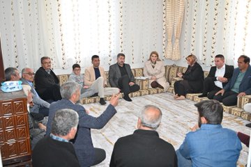 First image shows a group of about 15-20 people mostly men in suits and traditional attire with some women seated on floor cushions around a patterned rug in a room with beige curtains and wooden furniture one man in blue shirt gestures with hand raised during discussion. Second image depicts similar group of men and women in formal clothing sitting on cushions on a rug in a light-filled room with white curtains and a lamp some hold notebooks one man in gray suit points forward. Third image features a group of around 10 people including men in suits and a blonde woman in beige trench coat standing outdoors near white vehicles and tents on gravel under cloudy sky with autumn trees some smile and gesture. Fourth image shows similar outdoor group with men in various coats and the blonde woman in trench coat standing together near cars and tents on dirt ground engaging in conversation.