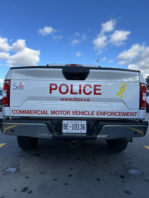 Rear view of a white Ford F-150 police pickup truck with DRPS Commercial Motor Vehicle Enforcement logos on the tailgate and doors yellow ribbon decals on the sides and tailgate Ontario license plate BE-10136 visible on the bumper and DRPS website URL printed below the enforcement text under a clear blue sky with scattered clouds in a parking lot setting.