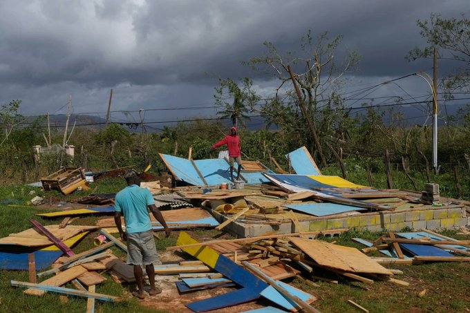 Two men in casual clothing stand amid scattered wooden and metal debris from destroyed structures on a rural plot with uprooted trees cloudy sky and utility poles in background one man wears blue shirt other red shirt surrounded by colorful sheet fragments and building remnants.