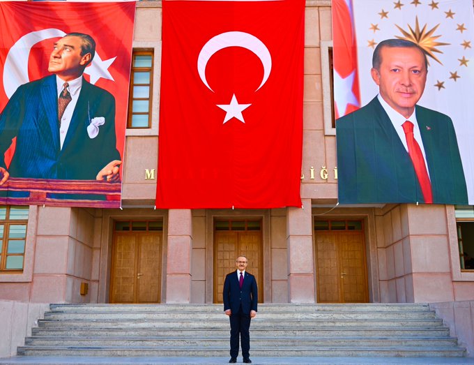 Large portraits of Recep Tayyip Erdoğan one in a dark suit with red tie gesturing forward and another in a green suit with red tie standing at a podium are displayed on either side of a massive red Turkish flag with white crescent and star hanging from a building facade. A man in a dark suit and tie stands centered on the steps leading to the buildings wooden double doors. Additional Turkish flags wave on the sides and the building shows architectural details like stonework and windows.