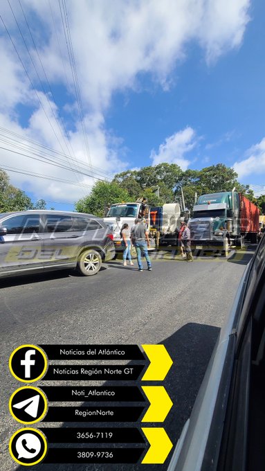 Two roadside images depict a traffic collision on a highway with clear blue skies scattered clouds and green trees in the background. A silver car is parked on the asphalt near two white trucks one with a crane arm extended and workers in casual clothing standing around discussing the incident. A red circle highlights a man in a gray shirt near the truck possibly the focused individual. Utility poles and wires run alongside the road with no visible damage to vehicles beyond positioning. Yellow promotional banners for Noticias del Atlantico appear in the lower corners including Facebook and phone contact icons.