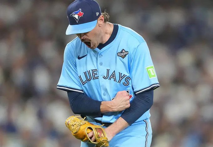 A baseball player wearing a light blue Toronto Blue Jays jersey with dark blue sleeves and pants, a matching cap, and a brown glove on his right hand, captured mid-pitch with his mouth open and right arm extended forward, standing on a baseball field.