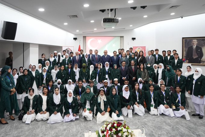 First image shows a large group of people including men and women in formal attire such as suits and hijabs seated on the floor in a conference room with a screen displaying Turkish and Pakistani flags a portrait on the wall and a flower arrangement in front. Second image depicts a diverse group of participants in traditional and modern clothing standing on grand stone steps of a historical building with ornate architecture and columns. Third image captures another group of individuals some wearing headscarves posing outdoors against a cityscape with a river bridge and holding Turkish and Pakistani flags.