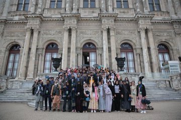 First image shows a large group of people including men and women in formal attire such as suits and hijabs seated on the floor in a conference room with a screen displaying Turkish and Pakistani flags a portrait on the wall and a flower arrangement in front. Second image depicts a diverse group of participants in traditional and modern clothing standing on grand stone steps of a historical building with ornate architecture and columns. Third image captures another group of individuals some wearing headscarves posing outdoors against a cityscape with a river bridge and holding Turkish and Pakistani flags.